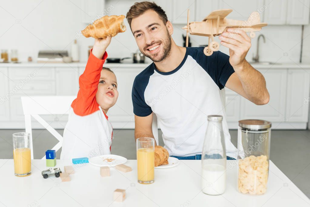 handsome young father and son playing with croissant and toy plane while having breakfast together at home