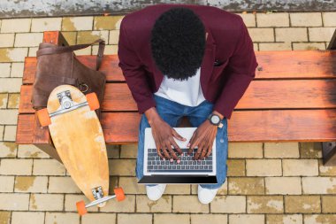 Genç Freelancer deri sırt çantası ve kaykay ile bankta dizüstü kullanarak üstten görünüm
