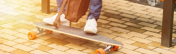 wide cropped shot of man with leather backpack and skateboard sitting on bench on street