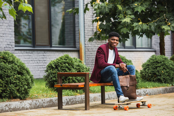 smiling young man with leather backpack and skateboard sitting on bench on street