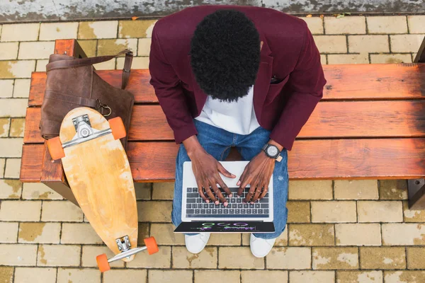 非洲裔美国人自由职业者使用笔记本电脑在板凳上的皮革背包和滑板的顶部视图 — 图库照片