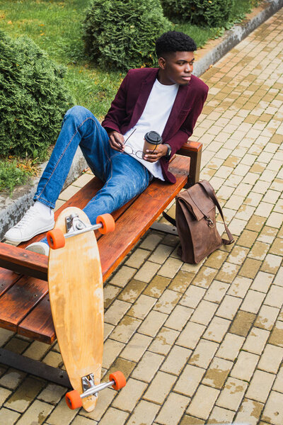 stylish young man with paper cup of coffee and eyeglasses sitting on bench