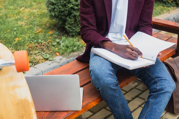 アフリカ系アメリカ人学生のラップトップでベンチでノートに書くのショットをトリミング  — 無料ストックフォト