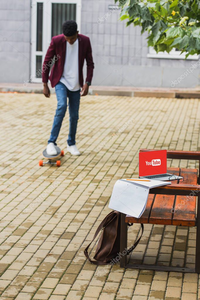 Young man riding skateboard on street with laptop with youtube logo on screen lying on bench on foreground