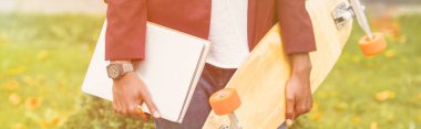 cropped wide shot of stylish student with notebooks and skateboard standing on street clipart