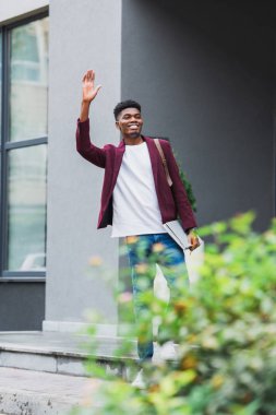 mutlu genç öğrenci sırt çantası ve sokakta el ile sallayarak dizüstü bilgisayarlar ile