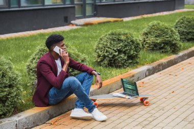 Genç serbest meslek sahibi kaykay üzerinde yalan ekranda sportsbet Web sitesi ile dizüstü bilgisayar ile sokakta kaldırım üzerinde otururken telefonla konuşuyor