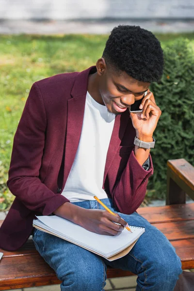 Sonriente Joven Estudiante Hablando Por Teléfono Escribiendo Cuaderno Sentado Banco — Foto de stock gratis