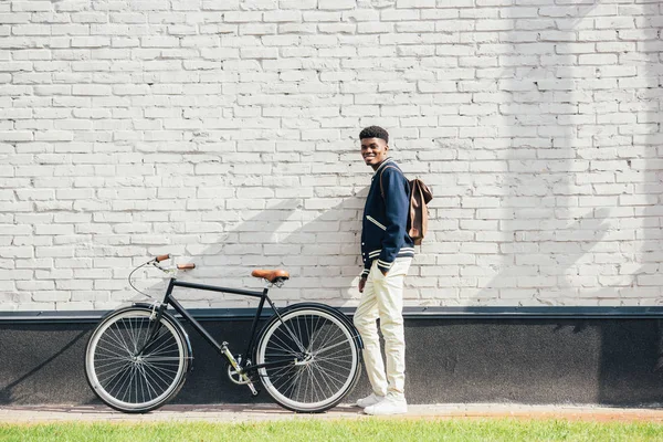 白いレンガの壁の近くの自転車で立っている革のバックパックと陽気なスタイリッシュなアフリカ系アメリカ人  — 無料ストックフォト