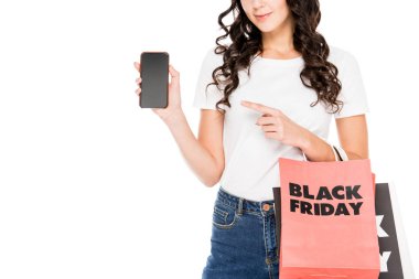 Kırpılan Smartphone cep telefonu ile boş bir ekran ile üzerine gelip alışveriş torbaları üzerinde beyaz izole siyah Cuma işaretiyle tutan kız görünümünü
