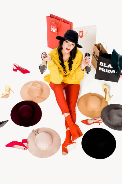 high angle view of young woman sitting on floor with shoes, hats and shopping bags with black friday lettering inscription around isolated on white