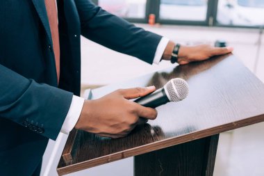 cropped image of african american lecturer standing at podium tribune with microphone in conference hall clipart