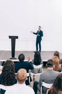 Seminer ve konferans salonunda ekranda işaret sırasında çok kültürlü kitleye konuşan öğretim görevlisi gülümseyen