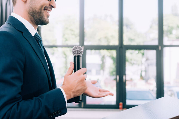 обрезанное изображение улыбающегося докладчика, жестикулирующего и разговаривающего в микрофон на трибуне подиума во время семинара в конференц-зале
