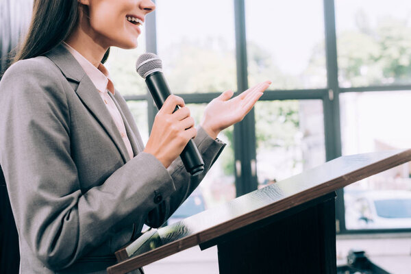 cropped image of smiling lecturer talking into microphone and gesturing at podium tribune during seminar in conference hall