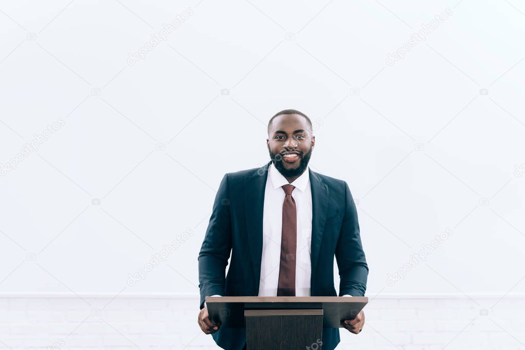 smiling handsome african american lecturer standing at podium tribune during seminar in conference hall and looking at camera