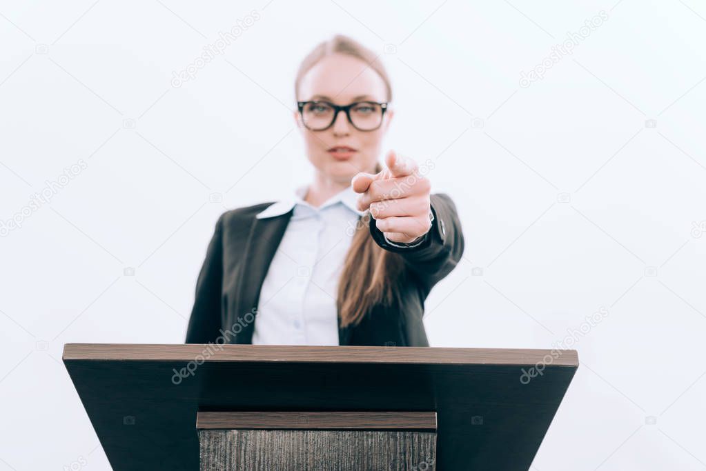selective focus of attractive lecturer pointing on camera from podium tribune isolated on white