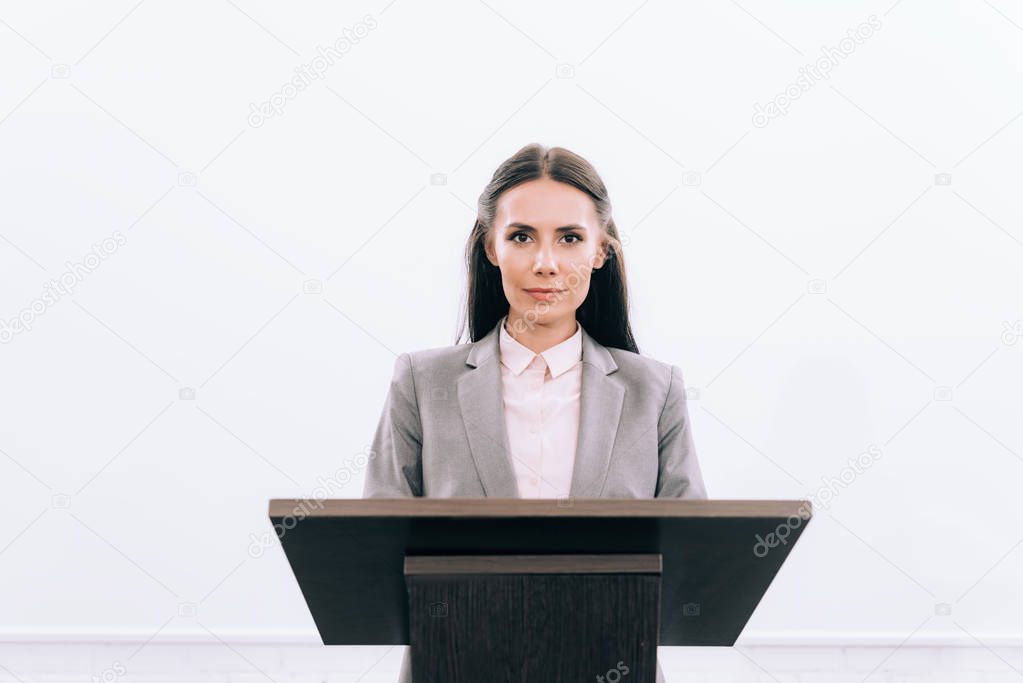 attractive lecturer standing at podium tribune during seminar in conference hall and looking at camera