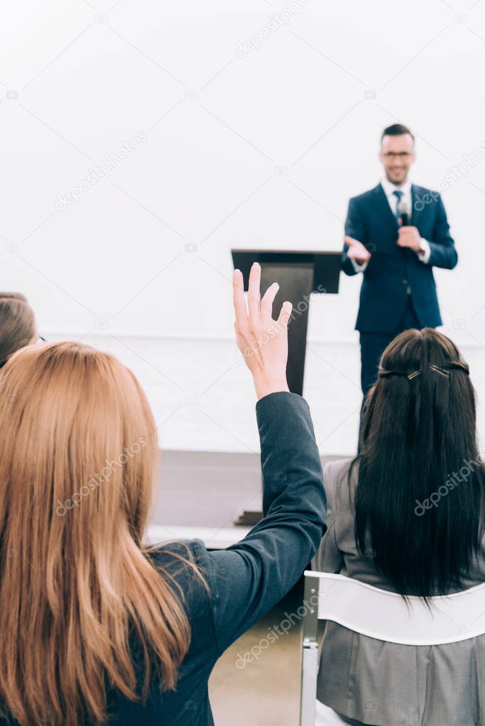 selective focus of lecturer talking in front of audience during seminar in conference hall, participant raising hand