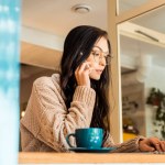 Hermosa mujer sentada en la cafetería con ordenador portátil y hablando por teléfono inteligente