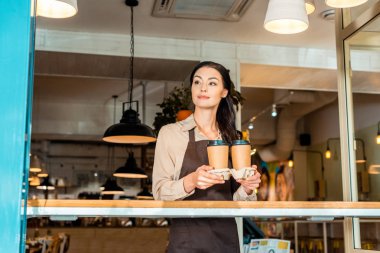 beautiful waitress in apron holding coffee in paper cups in cafe and looking away clipart