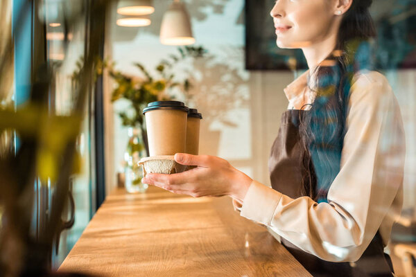 cropped image of waitress in apron holding coffee in paper cups in cafe