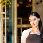 Portrait of beautiful waitress in apron looking at camera near cafe