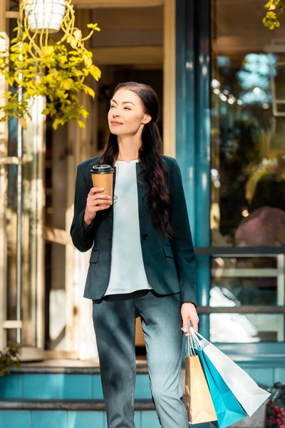 ショッピング バッグや使い捨てのコーヒー カップ カフェ近くの通りに立っている陽気な美人  — 無料ストックフォト