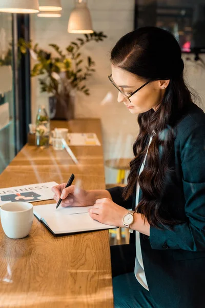 年轻的 Teleworker 写作在规划师在桌上咖啡和商业报纸在咖啡厅 — 图库照片