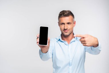 handsome adult man pointing at blank screen of smartphone isolated on white clipart