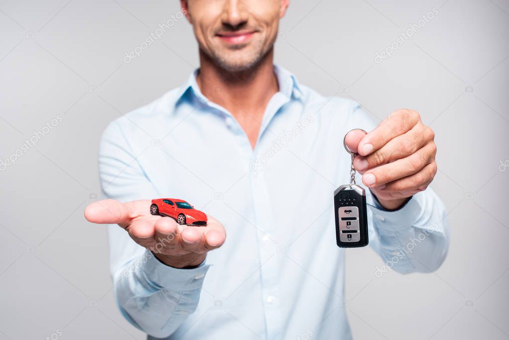 cropped shot of smiling adult man holding car alarm remote and toy red car isolated on white