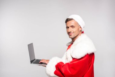 dizüstü bilgisayar kullanarak ve gri izole kamera gülümseyen santa kostüm yakışıklı adam 