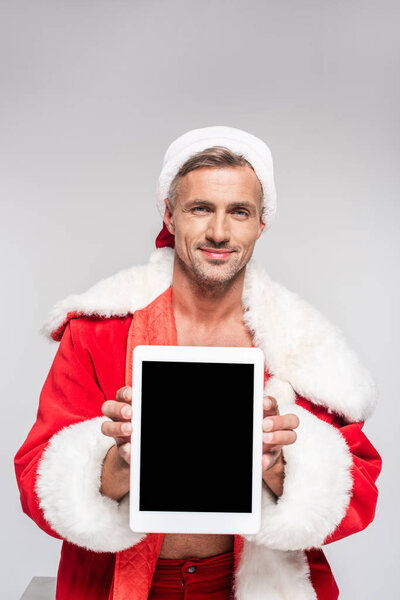 handsome man in santa costume holding digital tablet with blank screen and smiling at camera isolated on grey  