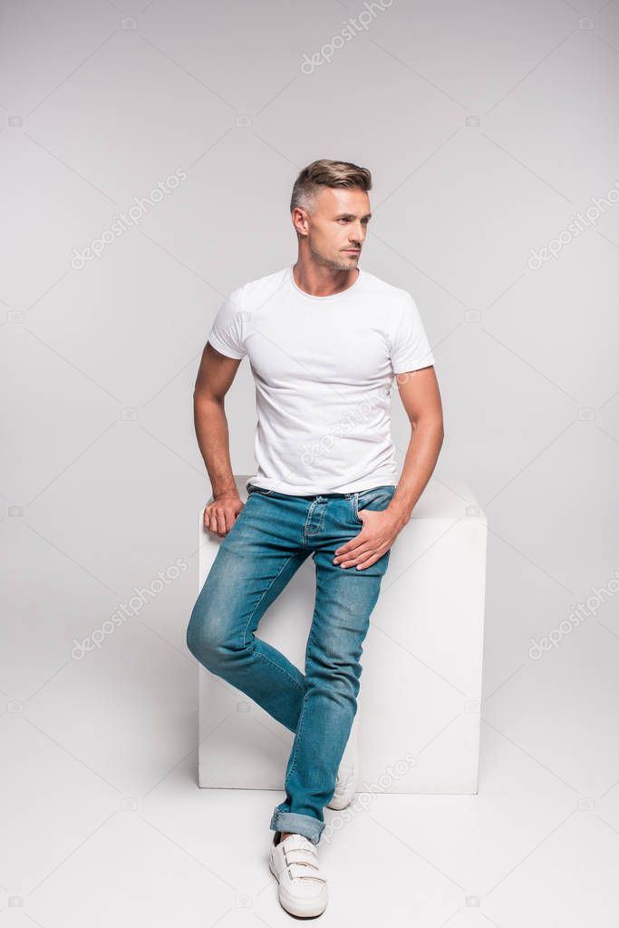 handsome man in white t-shirt and denim pants sitting and looking away on grey