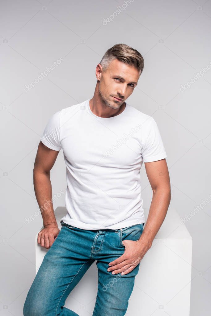 Portrait of handsome man in denim pants and white t-shirt sitting and looking at camera isolated on grey