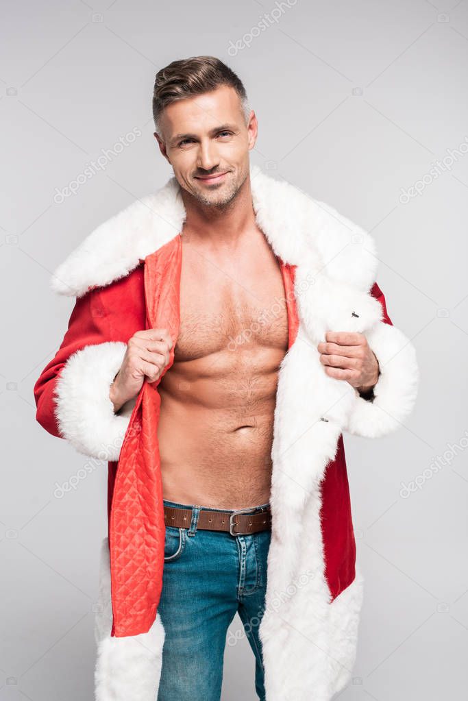 handsome muscular man wearing santa costume and smiling at camera isolated on grey 