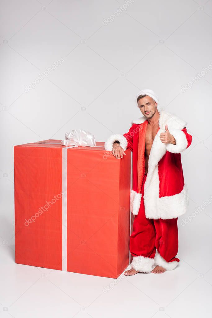 handsome santa claus leaning at big gift box and showing thumb up on grey