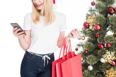 Kırpılan santa claus şapka ile alışveriş torbaları Noel ağacı üzerinde beyaz izole yakınındaki smartphone kullanarak kadında doz