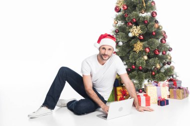 Noel ağacı üzerinde beyaz izole yakın oturan dizüstü bilgisayar ile Noel Baba şapkalı adam
