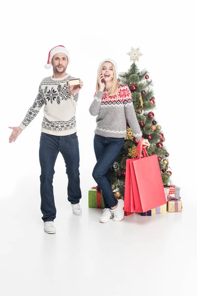 セーターや白で隔離クリスマス ツリー近く買い物袋でサンタ クロースの帽子のカップル  — 無料ストックフォト