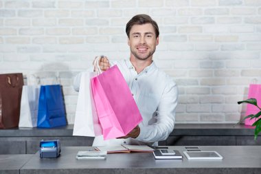 handsome young salesman holding shopping bags and smiling at camera in store clipart