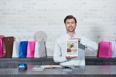 handsome young salesman holding digital tablet with ebay application and smiling at camera in shop  clipart