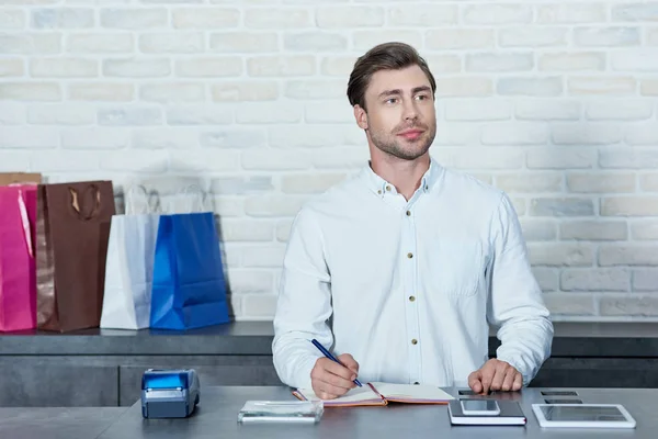 Young Man Writing Notebook Looking Away While Working Shop — Stock Photo, Image
