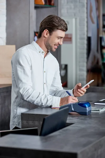 Uomo Sorridente Che Utilizza Smartphone Prende Appunti Mentre Lavora Negozio — Foto Stock