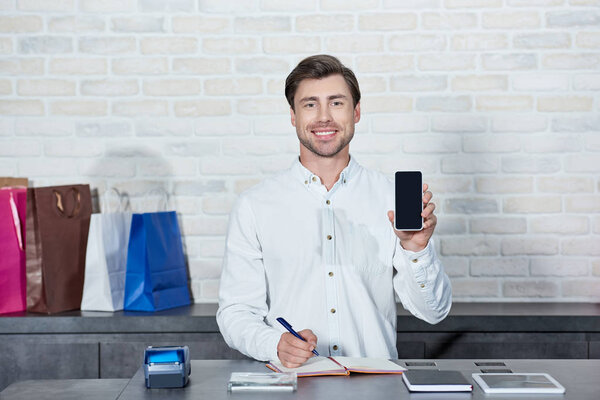 handsome young salesman holding smartphone with blank screen and smiling at camera in shop
