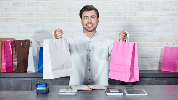 handsome young salesman holding shopping bags and smiling at camera in shop 