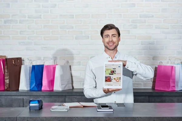 英俊年轻的推销员持有数字平板电脑与易趣应用程序和微笑在商店的相机 — 图库照片