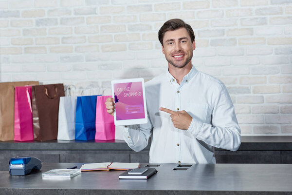 handsome young salesman pointing at digital tablet with online shopping application and smiling at camera in store   