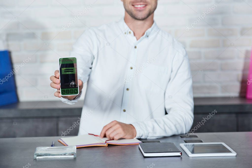 cropped shot of smiling young salesman holding smartphone with booking application in shop  