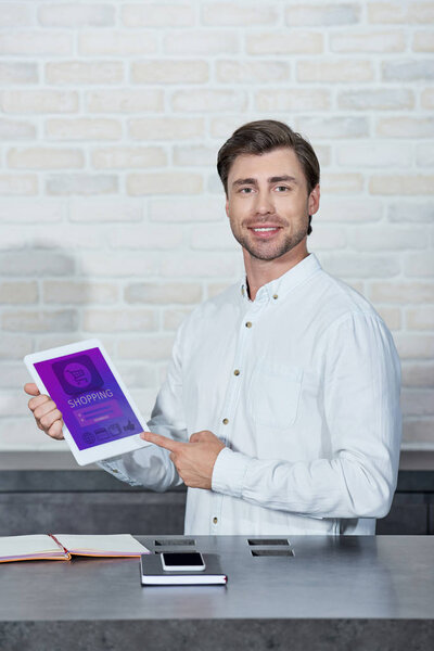 handsome young salesman holding digital tablet with shopping app and smiling at camera in store  